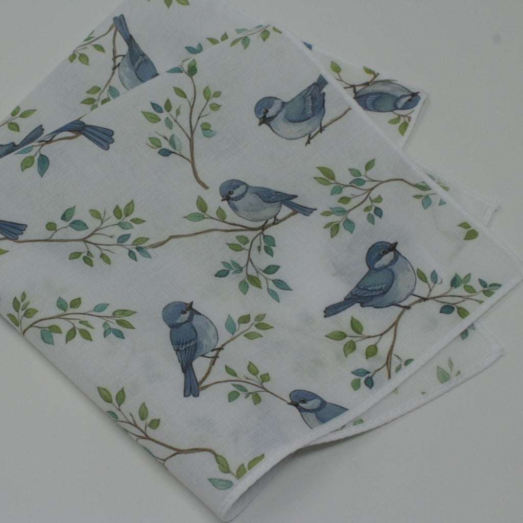 Cotton handkierchif A folded white fabric napkin or cloth featuring a printed pattern of blue birds perched on green and brown branches, displayed on a light gray surface.