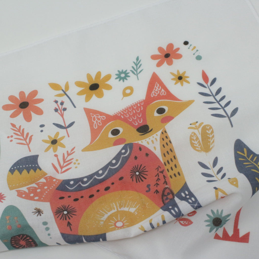 Cotton fabric handkerchief. A close-up of a white fabric featuring a colorful, whimsical illustration of a fox surrounded by flowers and leaves in orange, yellow, navy, and red.